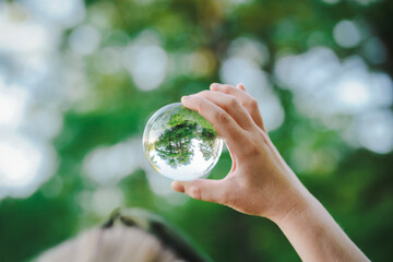 hands holding transparent glass ball or  crystal who reflects trees of a forest outside in green nature sphere symbolizing save the earth or planet