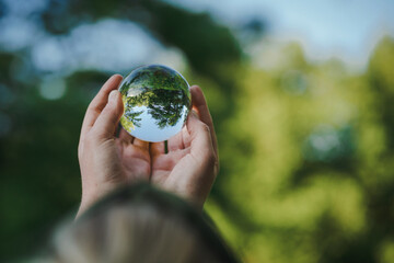 hands holding transparent glass ball or  crystal who reflects trees of a forest outside in green...