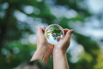 hands holding transparent glass ball or  crystal who reflects trees of a forest outside in green nature sphere symbolizing save the earth or planet