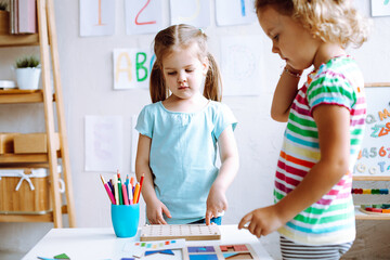 Two concentrated cute little girls children pupils standing at white table, playing board games in bright classroom.