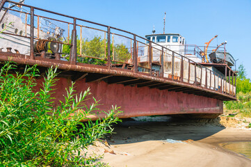 Obraz premium Old rusty ships lie on the sandy shore. Abandoned ships against a background of green bushes and blue sky. A rusting tugboat on the shore. Vessels that have served their service life. 