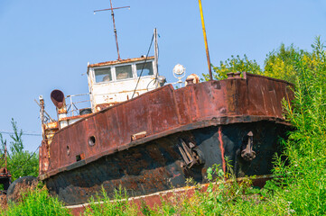 Old rusty ships lie on the sandy shore. Abandoned ships against a background of green bushes and blue sky. A rusting tugboat on the shore. Vessels that have served their service life. 