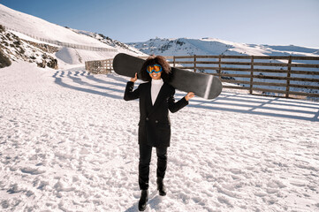 African American woman with goggles and a snowboard on a snowy mountain during winter
