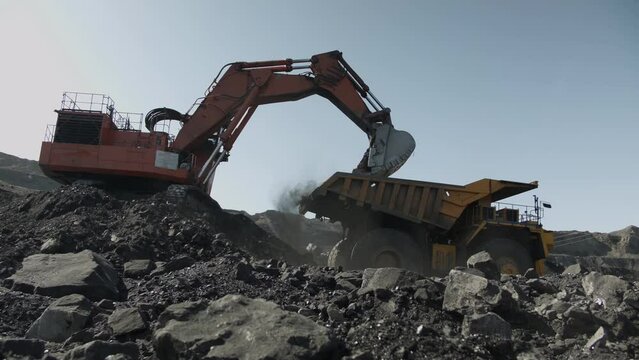 Excavator lifts bucket filled with coal and pours it into mining truck. Process of loading minerals in open pit