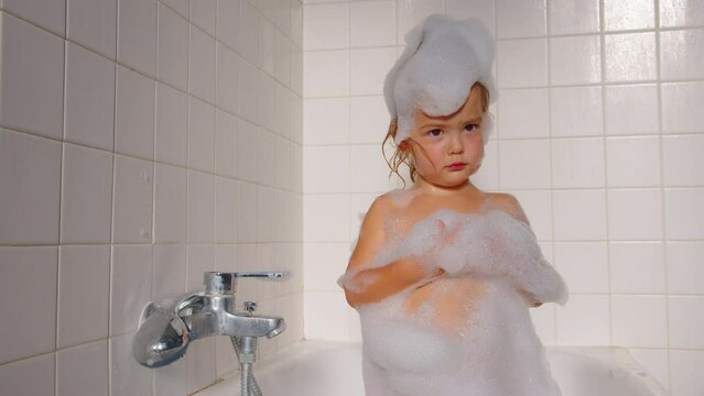 Close-up. Little cute girl bathes in a bath with foam. A child with foam on his head takes a shower. Attractive baby is basking in the bath. Daily hygiene and skin care for children.