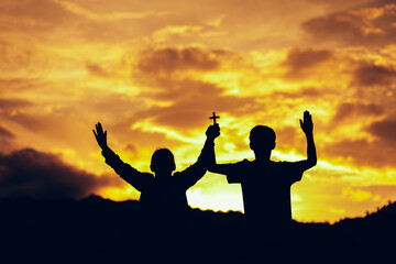 Fototapeta na wymiar Silhouette of two people holding cross together and praying worship to God at sunset. Hands in prayer. Christian Religion concept background. Copy space for your individual text.