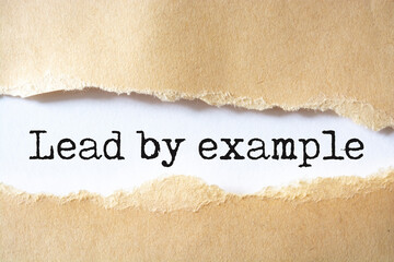 The text Lead by example appearing behind torn brown paper