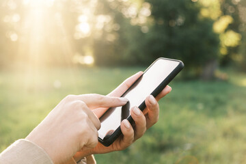 Closeup of female hands using a smart phone, nature background. Woman using mobile phone in sunset outdoors. Unrecognizable person touching smartphone screen. Unknown girl holding cellphone outside