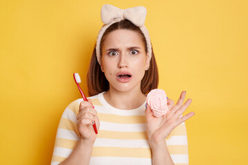 Obraz na płótnie Canvas Indoor shot of shocked amazed young woman wearing striped shirt and hair band posing isolated over yellow background, holding toothbrush and tasty marshmallow.
