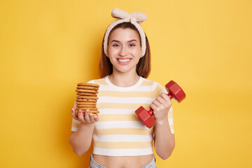Extremely happy young dark haired woman wearing striped shirt and hair band holding red dumbbell...