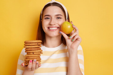 Attractive slip happy smiling woman wearing striped shirt and hair band, holding cookies and apple,...