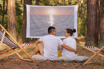 Outdoor shot of lovely young couple sitting in the forest in front of projector wall and watching...