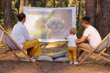Portrait of happy family sitting in the forest on deck chairs with their little daughter and watching movie on projector, parents playing with their cute kid and enjoying nature and fresh air.