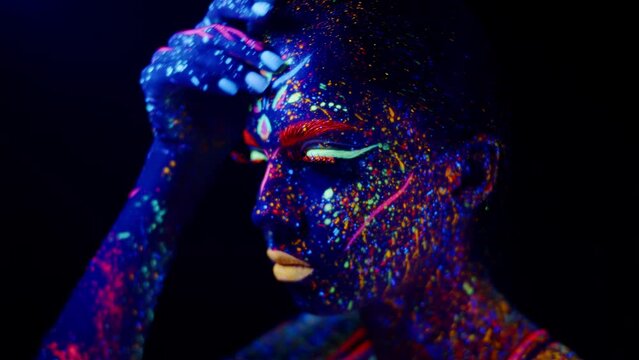 Woman in neon party makeup glowing in ultraviolet light touching her face, looking at her hand. Closeup portrait, recreational drug use, LSD acid trip concept.