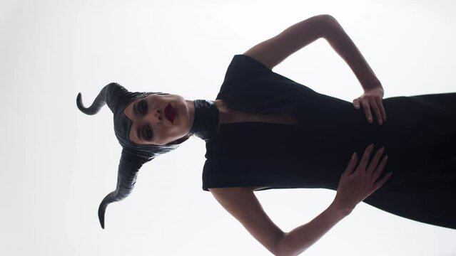 Attractive caucasian lady with horns in black clothing, posing with hands on her hips, looking into camera. Seductive evil demonic woman from a fairytale movie. Vertical video on a white background