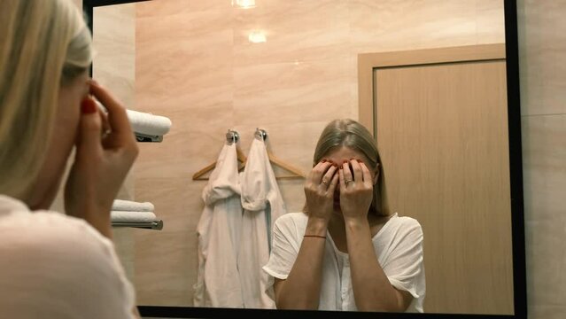 Ageing. Sagging skin. A young woman does facelift exercises by looking in the bathroom mirror. Facebuilding. Modern care, relaxation of facial muscles. Facelift, plastic surgery