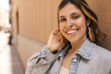 Beautiful young caucasian woman smiles, enjoys walk around city on sunny day. Brown-haired girl with piercing wears jacket in spring. Happy weekend concept.