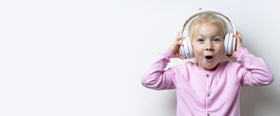 Surprised child girl in headphones looks up on a light background. Banner.