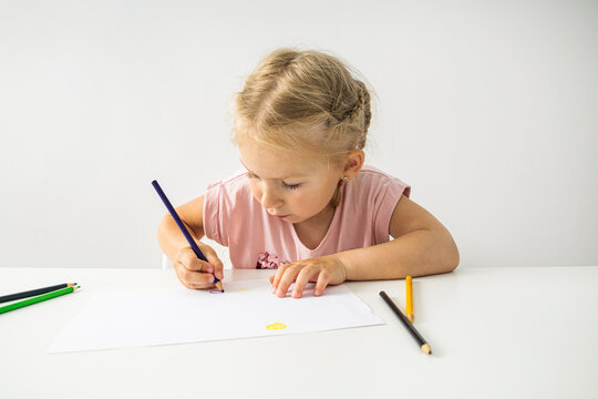 The child girl draws with colored pencils sitting at a white table on a white background
