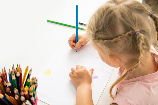 A child blonde girl draws with colored pencils sitting at the table. Top view, flat lay.