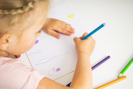 A child blonde girl draws with colored pencils sitting at the table