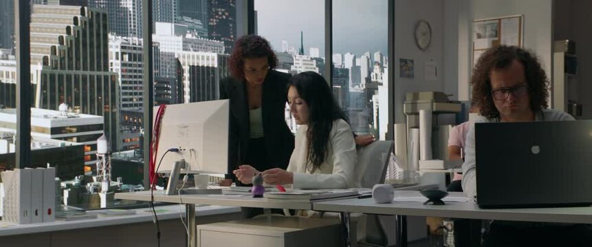 Two diverse female office employees working on something together in the modern busy office. View on a business district with skyscrapers in the background