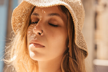 Close-up tender caucasian young lady with closed eyes is resting outdoors. Blonde girl wears piercing and hat on sunny day in summer. Leisure lifestyle and beauty concept.