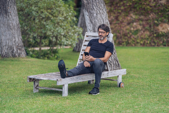 Middle-aged man sitting on a wooden hammock while using her mobile