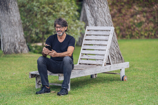 Middle-aged man sitting on a wooden hammock while using her mobile