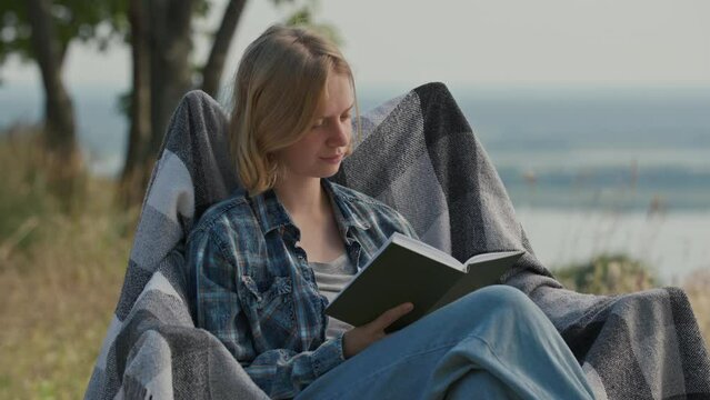 20s woman reading book sitting in tourist armchair outdoors. Young adult person reads paper book while camping time.