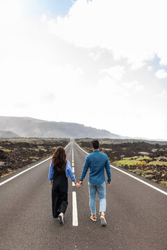 Anonymous young stylish couple holding hands and walking on road in highland