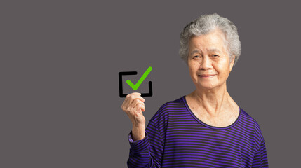 Senior woman holding a green check mark symbol looking at the camera with a smile while standing on...