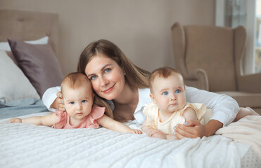 Tender portrait of smiling happy mother lying on bed with two little twin daughters 6 month old at comfortable home with pastel colors interior