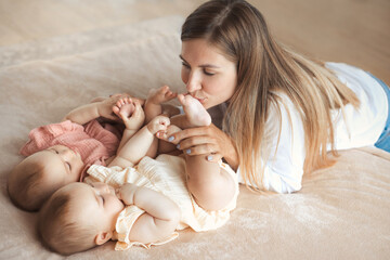 Young happy mother kissing barefoot legs of two baby twin sisters on the soft bed at home