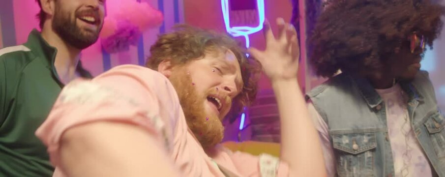 Young ecstatic man with confetti on beard and painted nails dancing and having fun at disco with friends in party room with neon light