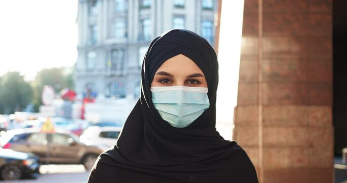 Young muslim Arab woman wearing hijab veiling face looking at camera outdoors during sunny day. Beautiful female Islam lady in medical mask covered with black headscarf. Religion, covid-19, virus.
