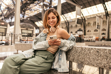 Fototapeta premium Nice caucasian young woman looks into smartphone, waiting for train sitting at station. Blonde girl wears T-shirt, pants and jacket. Concept of use