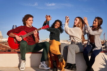 Cheerful women clapping hands near man playing guitar on terrace