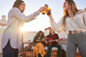 Smiling women clinking beer bottles near friends playing guitar