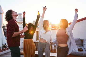 Delighted friends drinking beer and dancing on rooftop at sunset