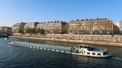 Obraz premium Transport fluvial de marchandises par péniche sur la Seine, dans la ville de Paris, avec des immeubles haussmanniens bordant le fleuve, quai de la Mégisserie (France)