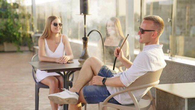 Relaxed man sitting in the company of two girls smoking hookah. Young people are chatting enjoying time in a cafй.
