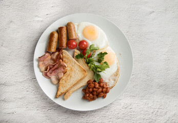 A white plate with scrambled eggs, bacon, beans, sausages, tomatoes and croutons on a light background.
