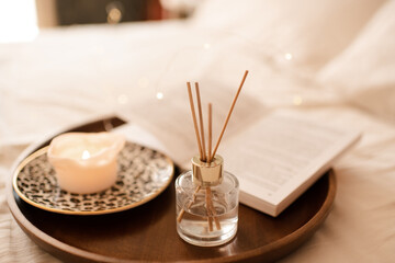 Aroma sticks with liquid scented perfume water in bottle with burning candle staying on open paper book on wooden tray in bed close up. Aromatherapy. Home cozy atmosphere.