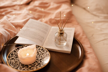 Aroma sticks with liquid scented perfume water in bottle with burning candle staying on open paper book on wooden tray in bed close up. Aromatherapy. Home cozy atmosphere.