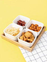 Four ceramic plates and a wooden tray on yellow background. Compartmental dish. Beer snacks. Sausages, crackers, nuts and dried squid