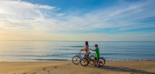 The lifestyle of a mother and son family on a bicycle at the sandy beach by the sea. The background image is a beautiful sunrise and sky. happy family concept