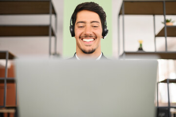Young latin businessman with headset sitting at the desk and talking by video with new customers or...