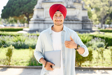 Positive smiling Indian man freelancer in turban showing thumbs up standing outdoors and holding...