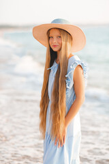 Beautiful smiling blonde teenager girl 12-14 year old with long hair wear straw hat and stylish elegant dress stand over sea coast outdoor. Summer season. Childhood.